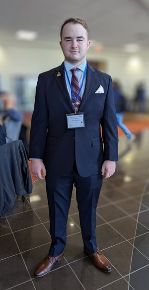 Benjamin Chicoria, the 2024 Roberts/Caperton Inspiration Award winner, dressed in a suit with a medal and name badge, standing in a lobby area.