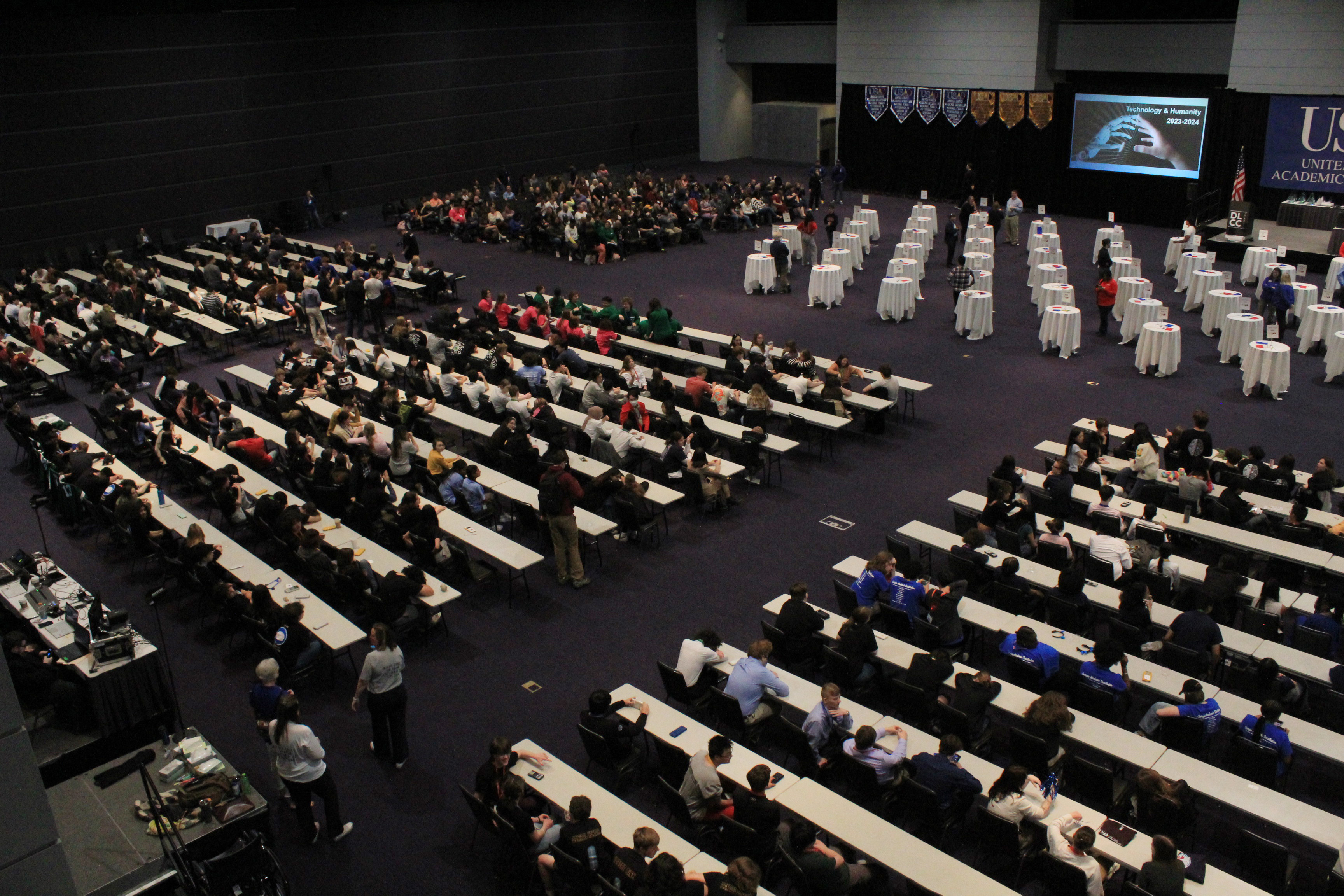 A large competition hall filled with United States Academic Decathlon (USAD) participants seated at long tables, preparing for an event. A stage with a screen displaying competition content is visible in the background. The image captures the intensity and focus of the competition experience.